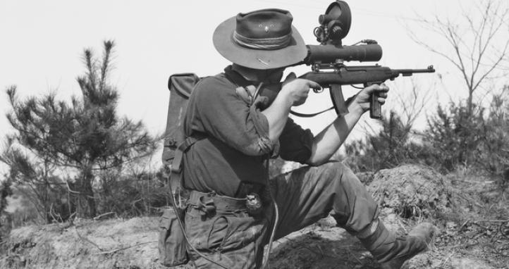 Private L. J displaying the Sniperscope which is attached to a .30 calibre Carbine (US Pat.) on 15 April 1952 in Kapyong Valley, Korea. The scope is battery operated and the battery is carried in the pack on his back.