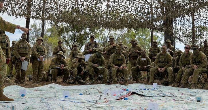 An Army officer addressing soldiers under camouflage netting and over a very large map on the ground.