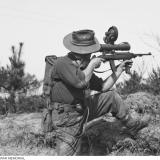 Private L. J displaying the Sniperscope which is attached to a .30 calibre Carbine (US Pat.) on 15 April 1952 in Kapyong Valley, Korea. The scope is battery operated and the battery is carried in the pack on his back.